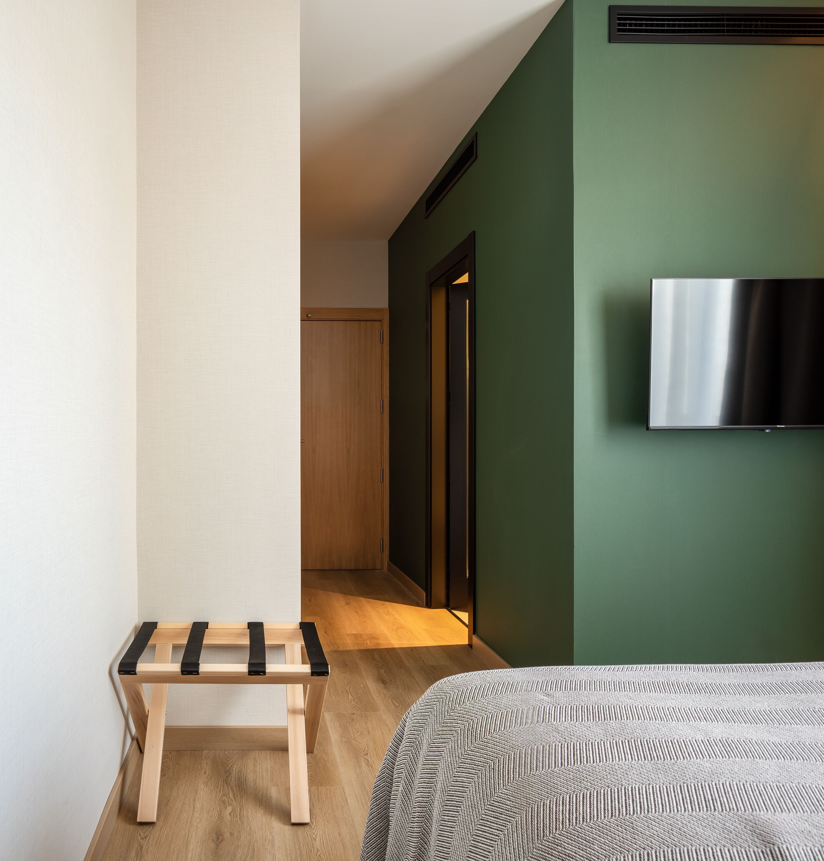 A hotel room with a bed, a small wooden bench, a television on the wall, and a door leading to another room.