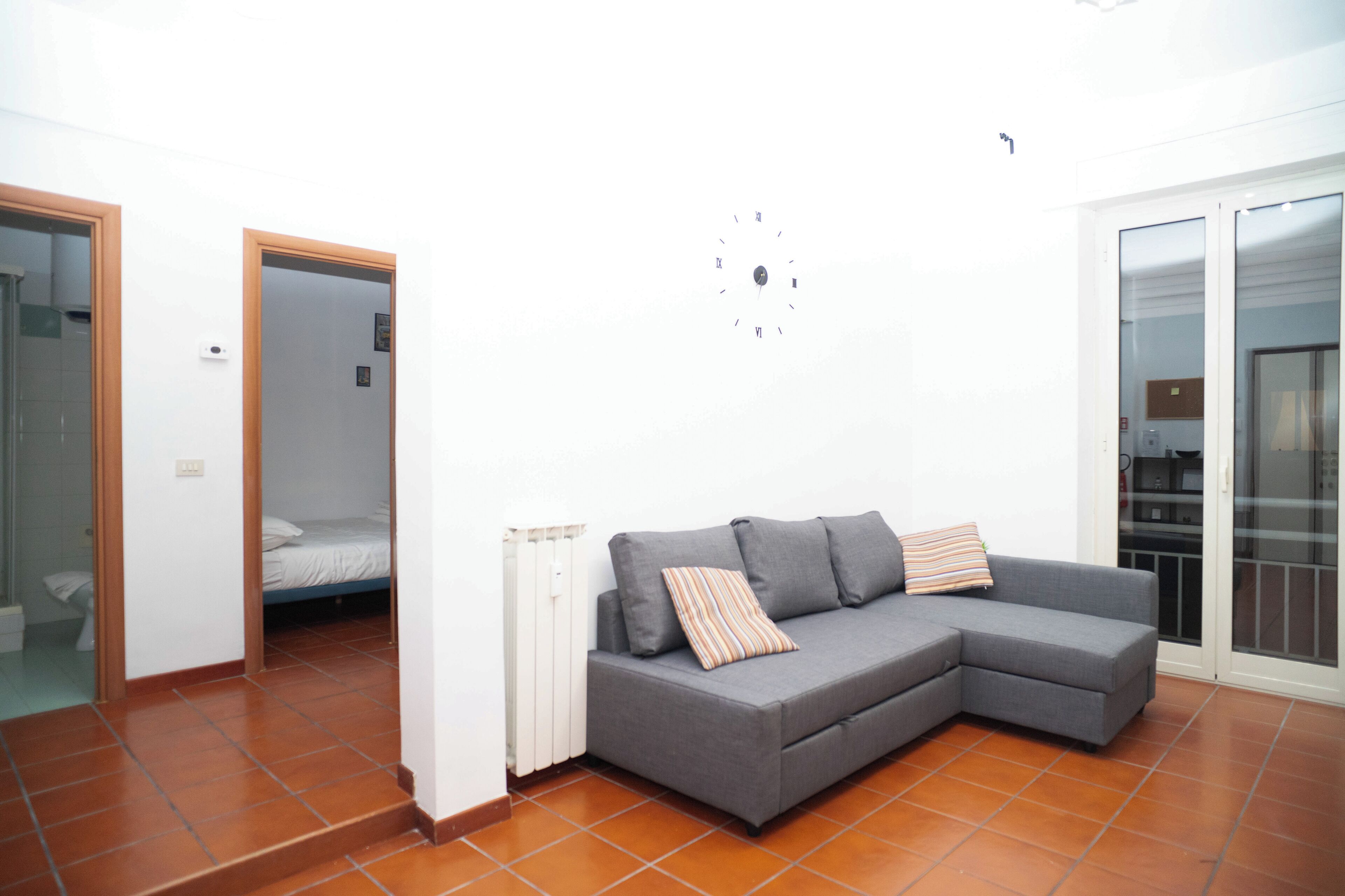 Una sala moderna con un sofá seccional grisáceo, una cama pequeña al fondo y un ventanal corredero que da a un balcón.