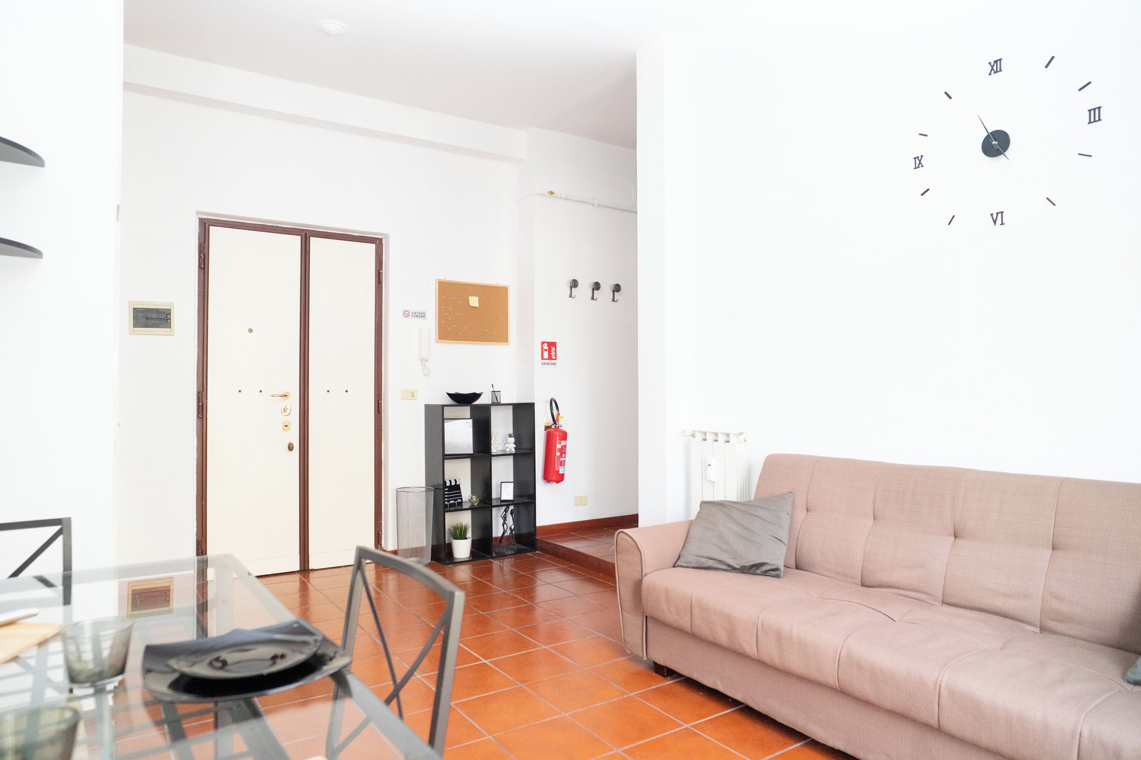 A living room with a sofa, a glass table, chairs, a clock on the wall, and a door in the background.