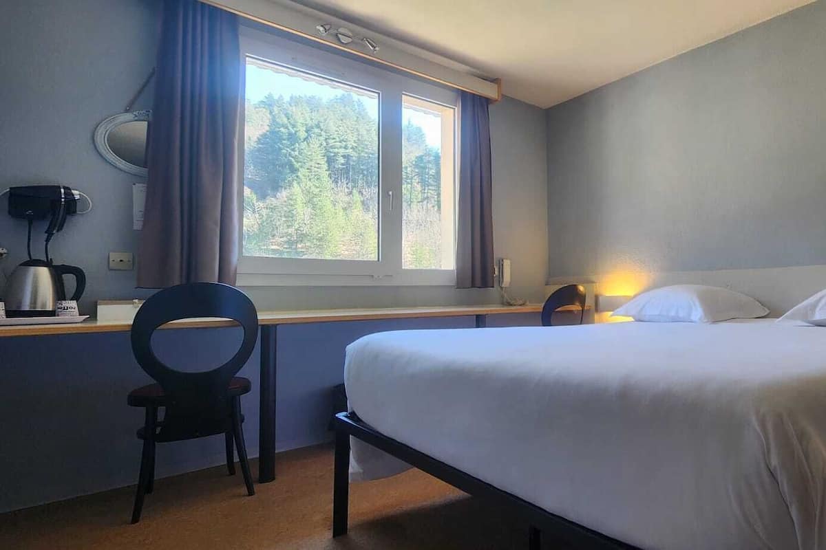 Un dormitori amb llit, escriptori amb cadira, finestra amb cortines i làmpada penjada a la paret.