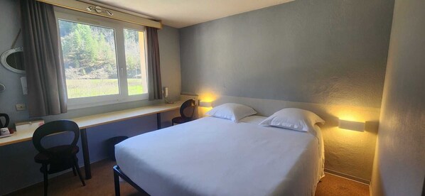 Une chambre d’hôtel équipée d’un lit, d’un bureau, d’une chaise, d’une fenêtre avec des rideaux et d’une lampe fixée au mur.