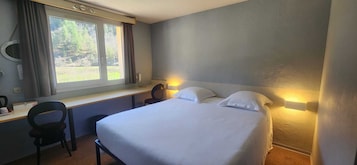 Quarto de hotel com cama, escrivaninha, cadeira, janela com cortinas e luminária na parede.