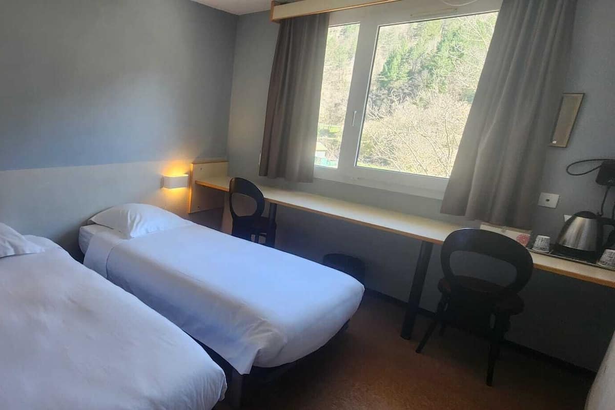 Habitació d'hotel petita i senzilla amb dos llits individuals, una taula de treball amb cadira, una finestra amb cortines i una làmpada penjada a la paret.