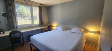 Quarto de hotel com cama, escrivaninha, cadeira e janela com cortinas.