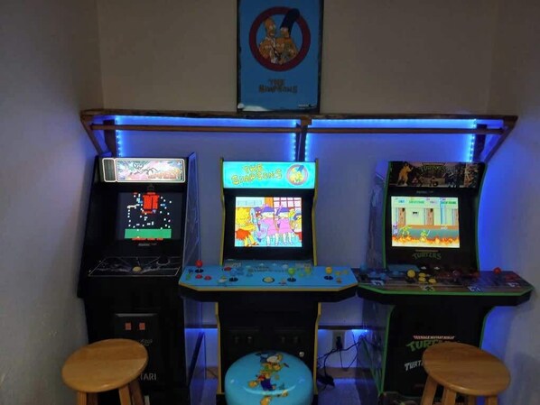 Three arcade machines with 'The Simpsons' and 'Turtles' games, a stool, and a 'No Smoking' sign on the wall.