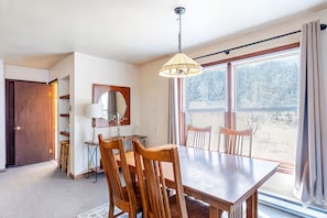 A dining area with a wooden table and chairs, a hanging lamp, a mirror, and a view of a building through large windows.