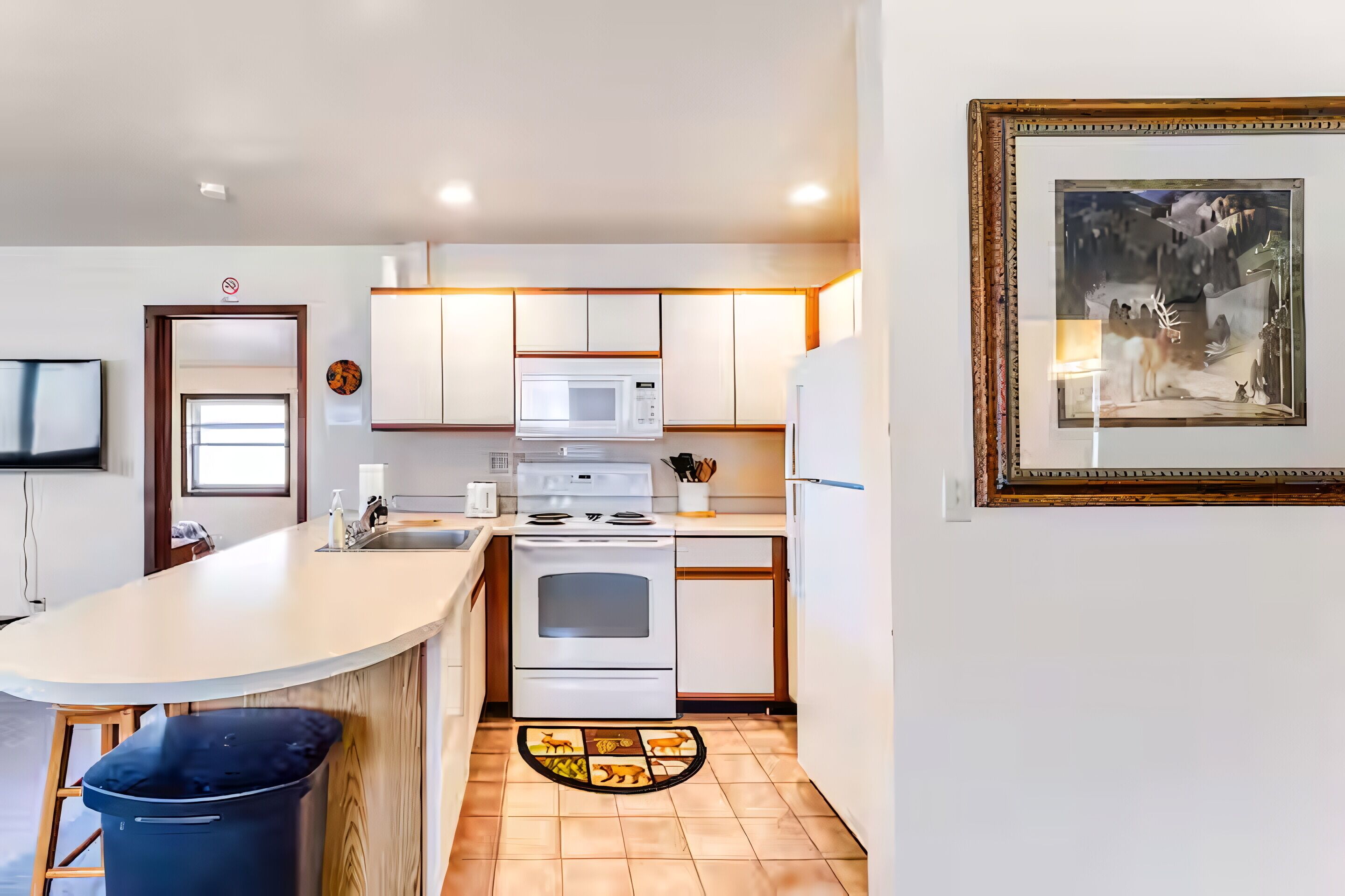 A modern kitchen with white cabinets, a microwave, stove, and a sink. There is a small island with a bar stool and a trash can. A framed picture hangs on the wall.