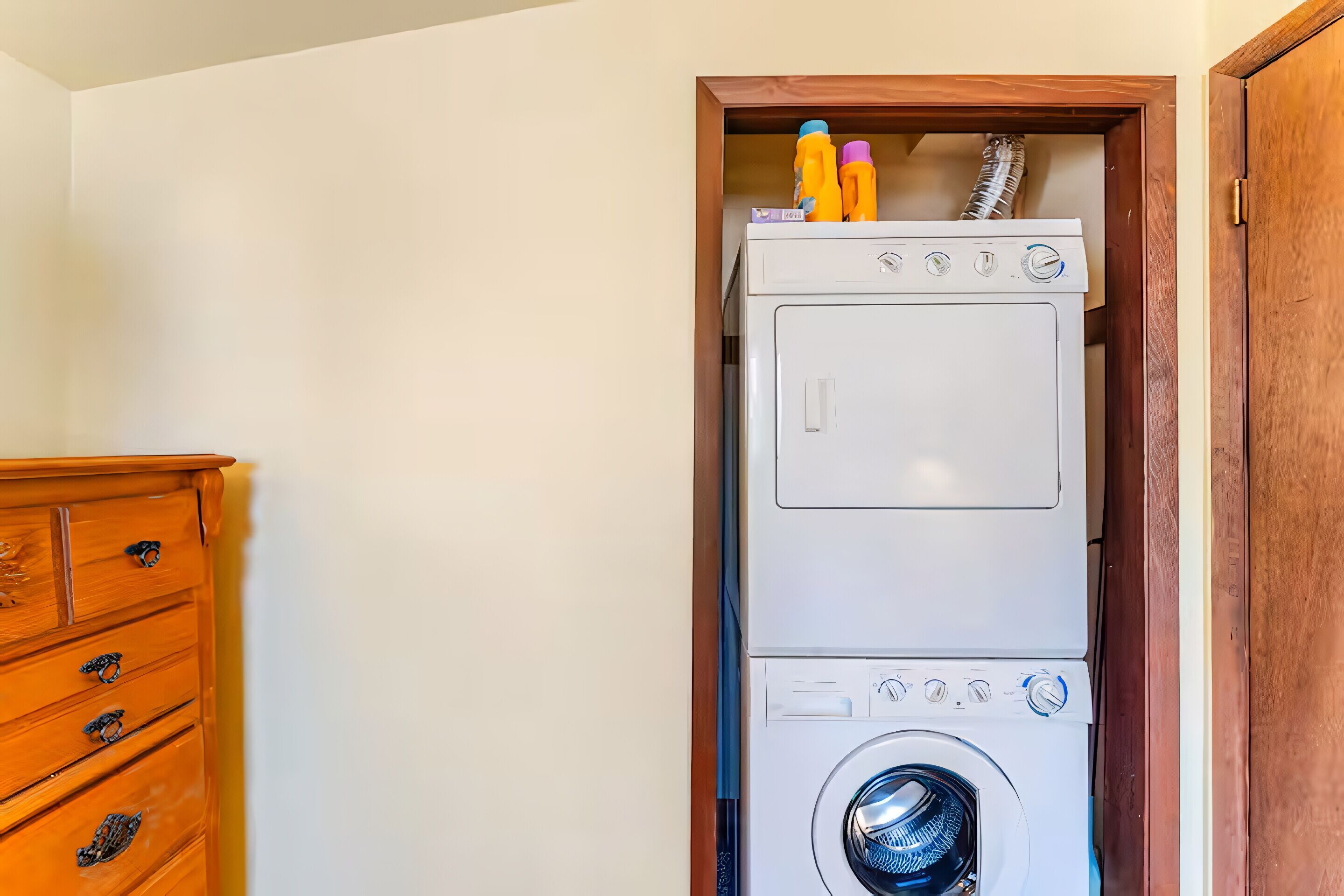 Un appareil compact, blanc, comprenant une laveuse et une sécheuse superposées, avec un meuble en bois placé à gauche.