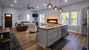 Uma cozinha moderna com ilha central, área de jantar com mesa e cadeiras, sala de estar com sofá e lareira, e janelas amplas que oferecem luz natural.