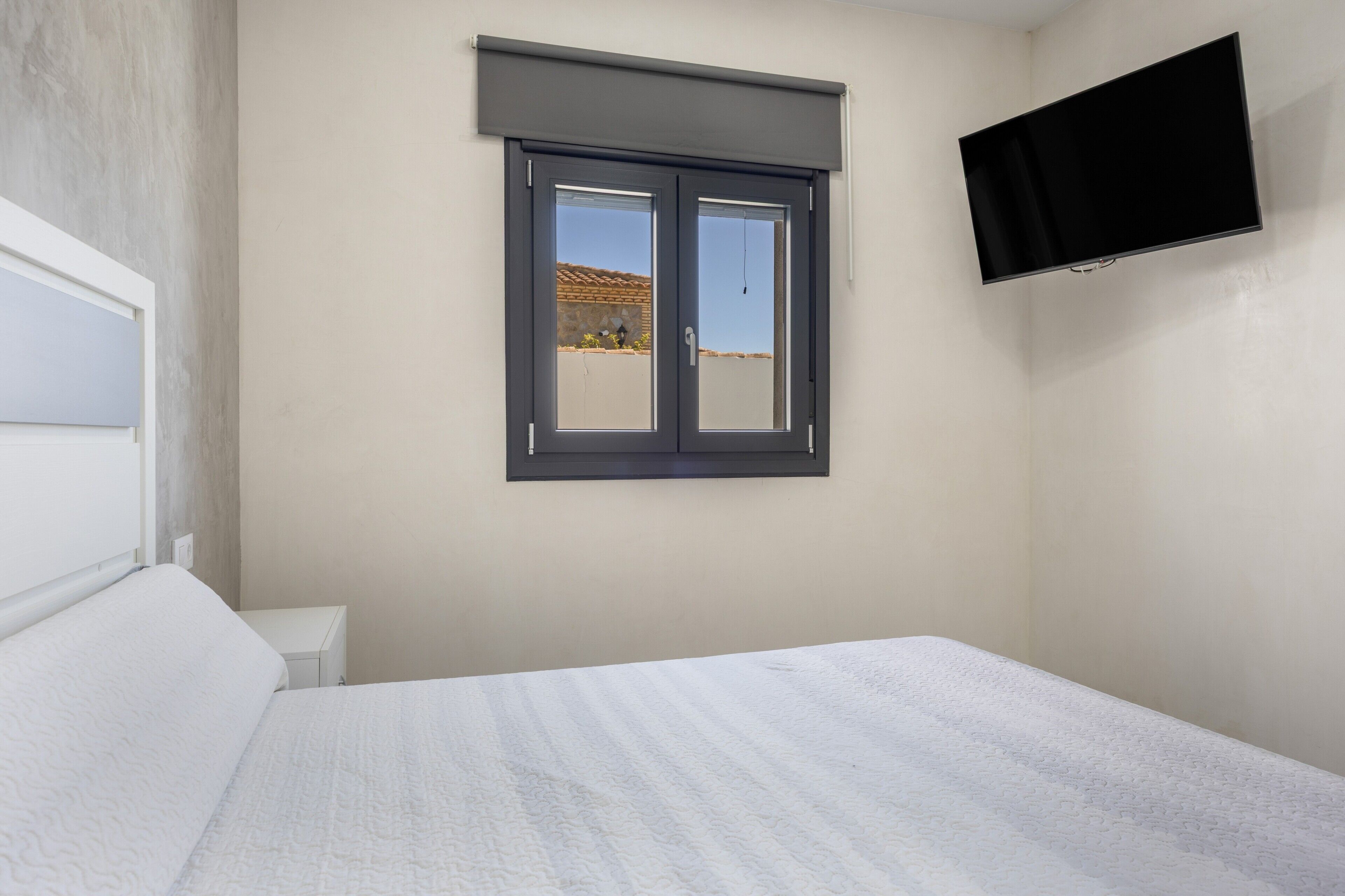 Une chambre à coucher avec un lit, une fenêtre avec des stores, un téléviseur à écran plat fixé au mur et une petite table de chevet.