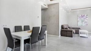 Una sala de estar moderna con una mesa de comedor blanca, cuatro sillas negras, un sofá gris y una mesa de centro blanca.