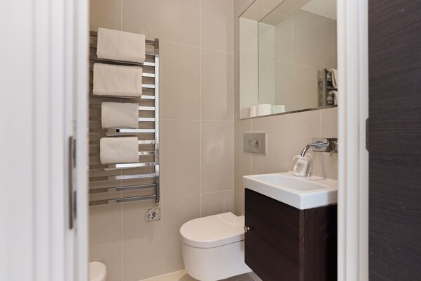 A modern bathroom with a toilet, a towel rack with multiple towels, a sink with a mirror, and a dark wooden cabinet. - Second Storey at Kew (Brentford)