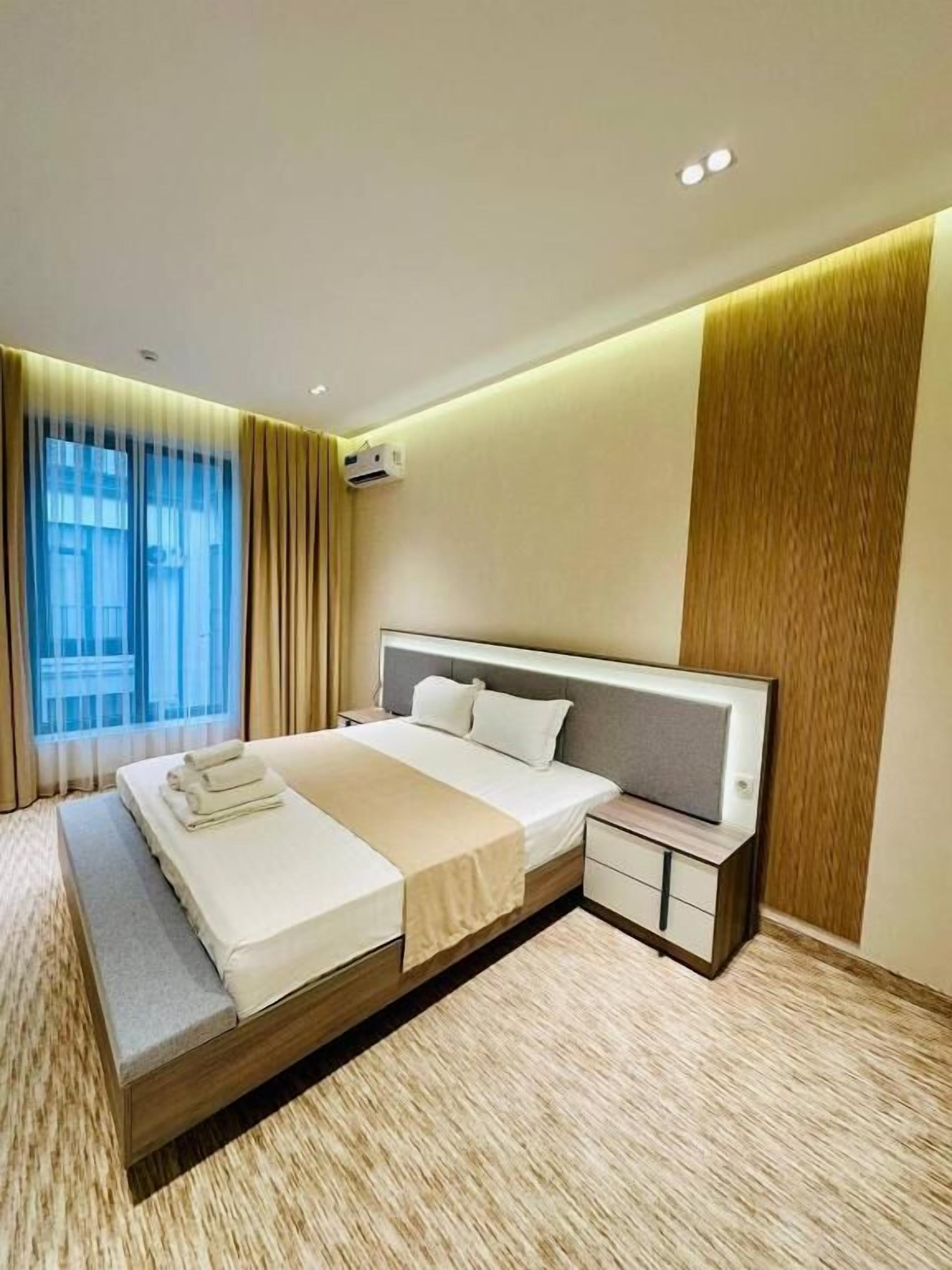 A modern hotel room with a large bed, a bedside table, an air conditioning unit, and a view of the city through the window.