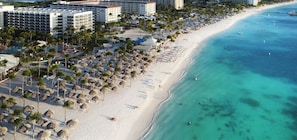 Ein Strandresort mit mehreren Hotelgebäuden, einem sandigen Strand, der von Palmen und Sonnenschirmen gesäumt wird, und klarem, türkisblauem Wasser.