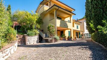 A two-story building with a stone staircase leading to the entrance, surrounded by greenery and a cobblestone driveway.