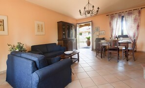 Un salon avec un canapé, une table à manger entourée de chaises, un lustre et une vue sur l’extérieur par une porte ouverte.
