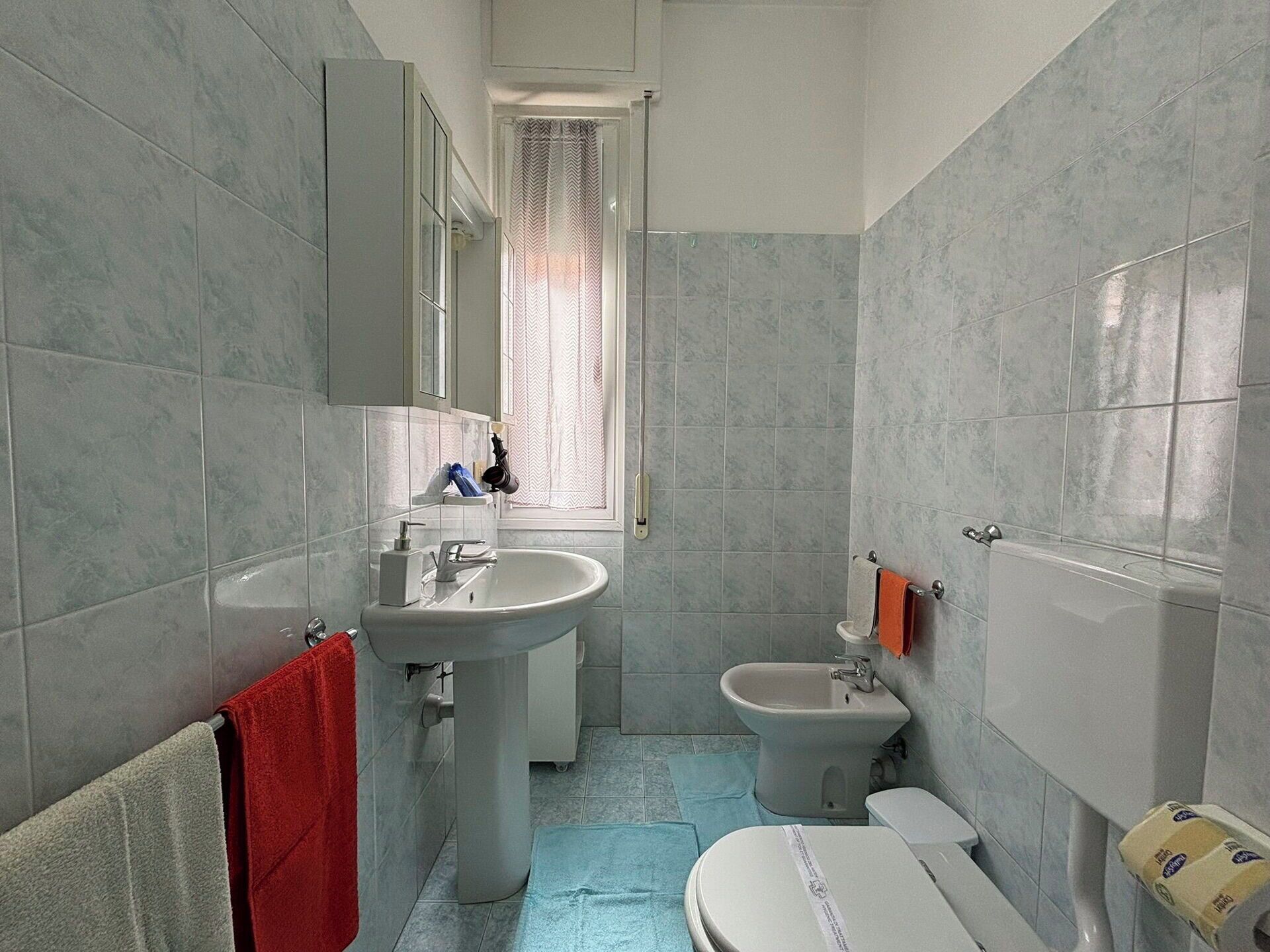 A bathroom with a sink, bidet, toilet, and a window with curtains.