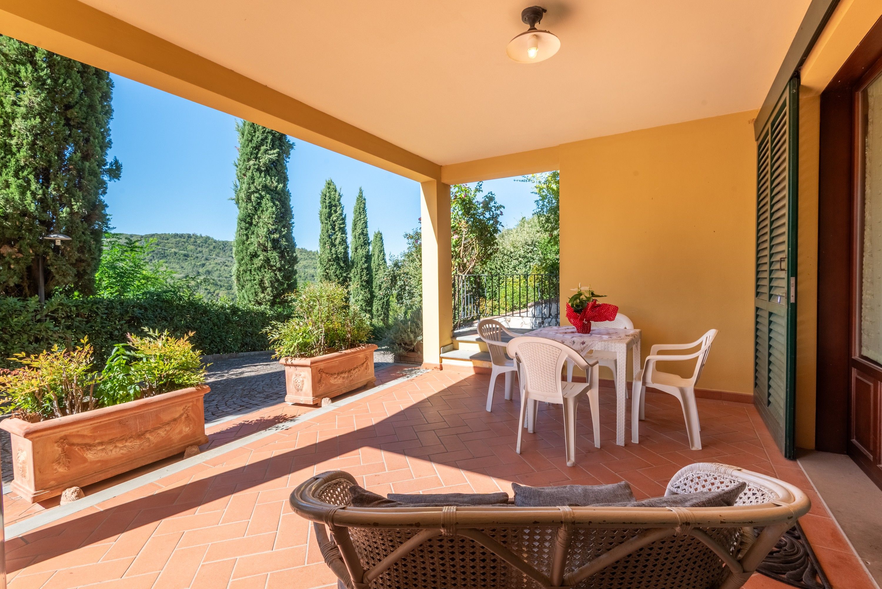 A covered patio with a table and chairs, potted plants, and a view of the surrounding landscape.