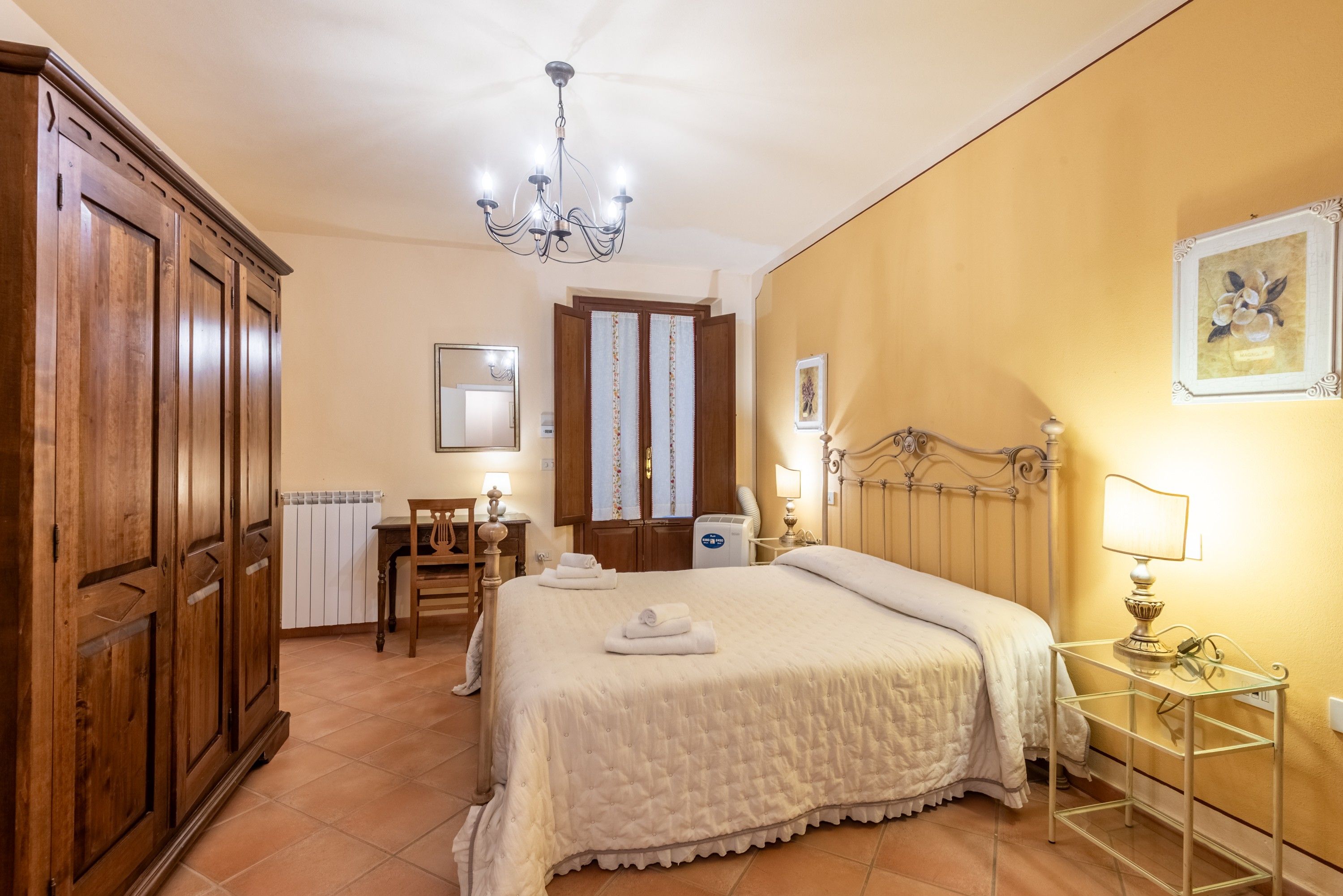 A hotel room with a bed, a desk, a chair, a wardrobe, and a chandelier.