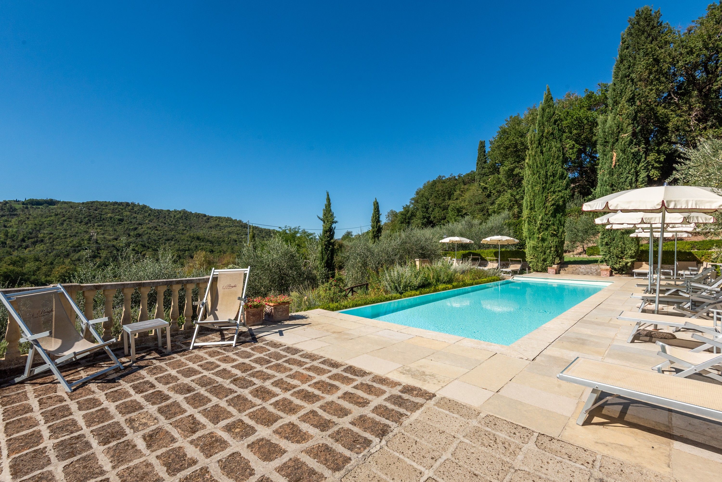 A pool area with lounge chairs and umbrellas, surrounded by greenery and hills.
