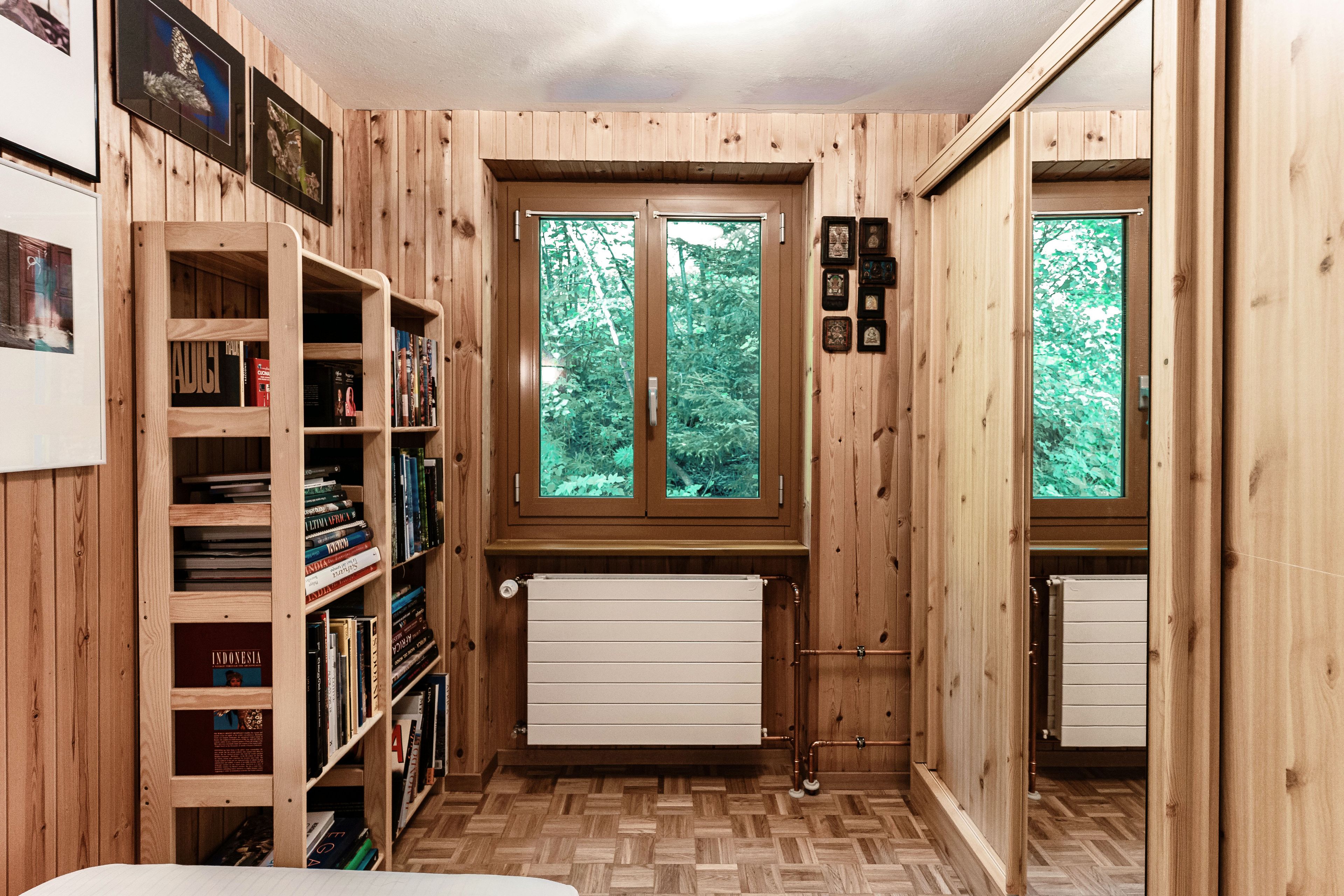 A wooden-paneled room with a bookshelf, radiator, and two windows with a view of greenery.