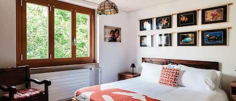 A bedroom with a bed, a chair, a nightstand, a lamp, and a window with a view of greenery.