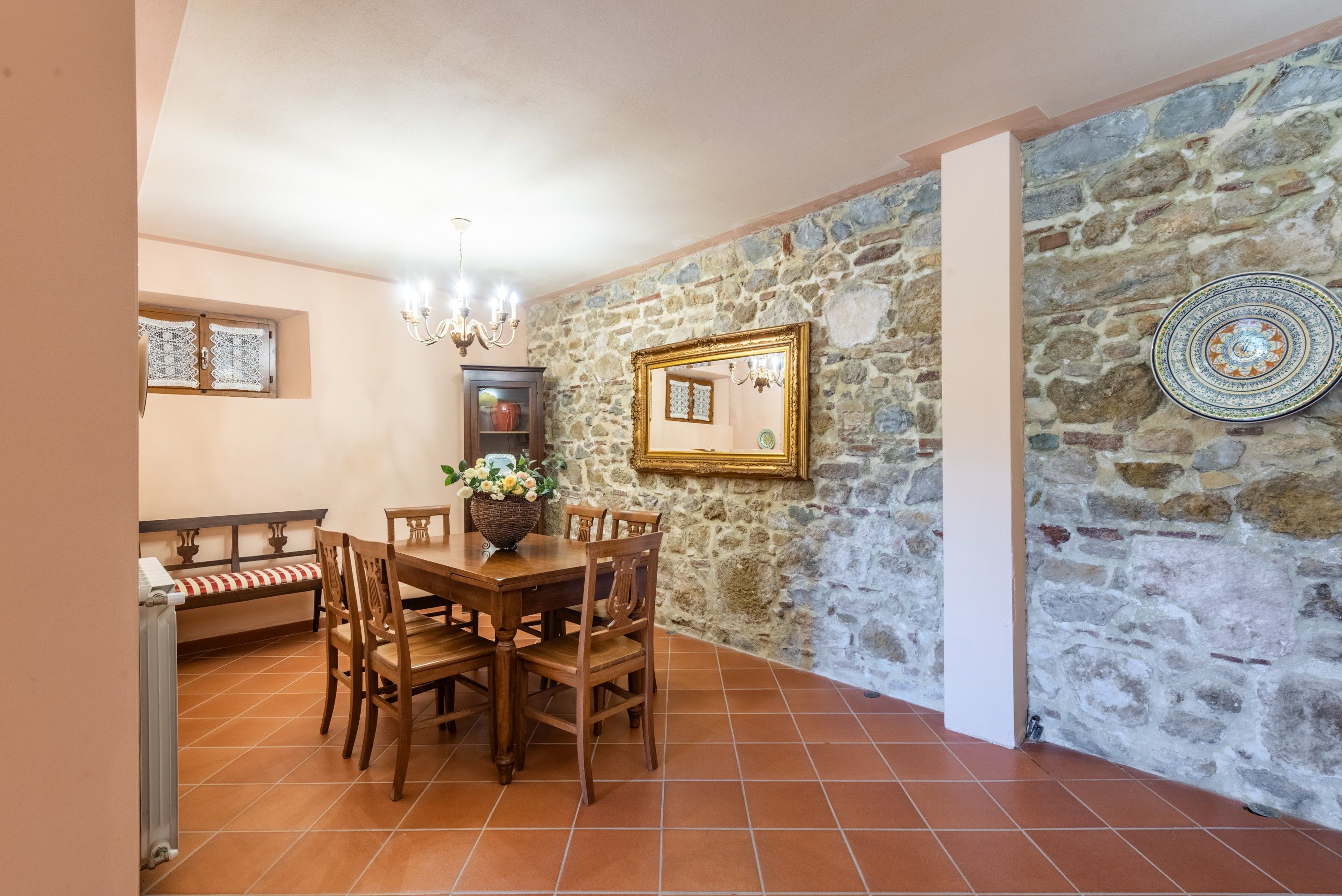 A dining area with a stone wall, a wooden table with chairs, a chandelier, and a cabinet with a lace cover.