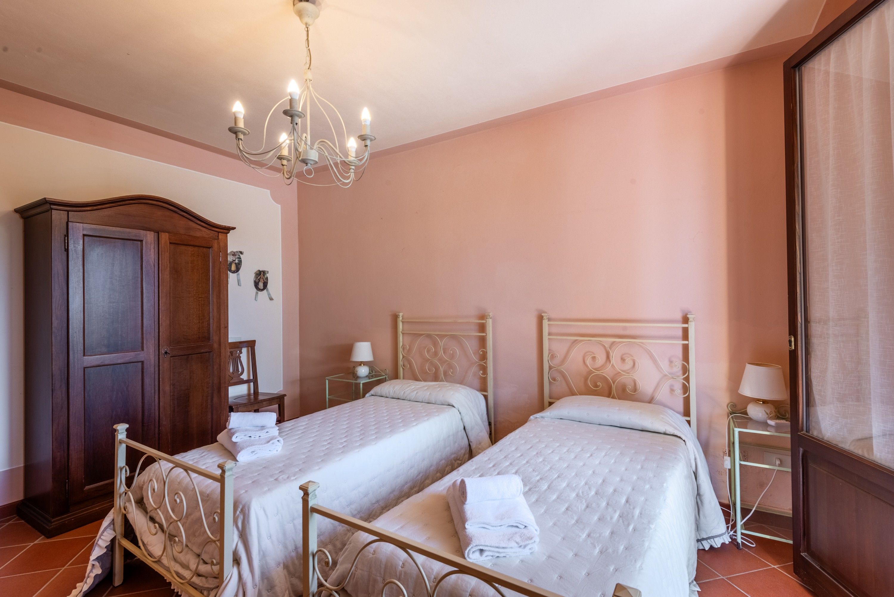A hotel room with two beds, a wooden wardrobe, a chandelier, and a window with curtains.