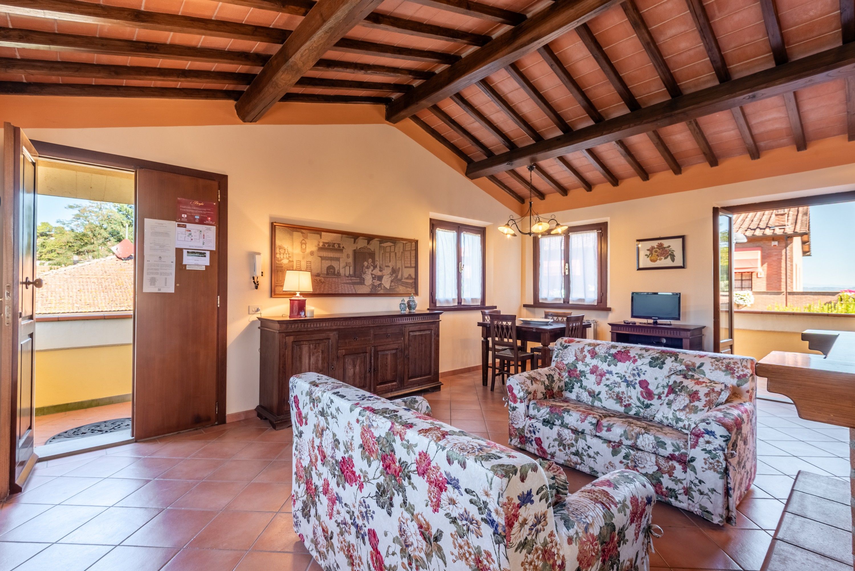 A spacious living area with a dining table, two floral sofas, a kitchen with a wooden ceiling, and a view of the outside.