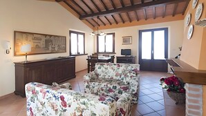A living room with a sofa, a dining table, a TV, and a kitchen area with a window. - Agriturismo I Borghi – Apartment Zenzero with Pool in San Gimignano (San Gimignano)