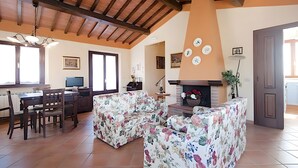 A living room with a fireplace, floral sofas, a dining area with a table and chairs, and a kitchen area with a stove. - Agriturismo I Borghi – Apartment Zenzero with Pool in San Gimignano (San Gimignano)