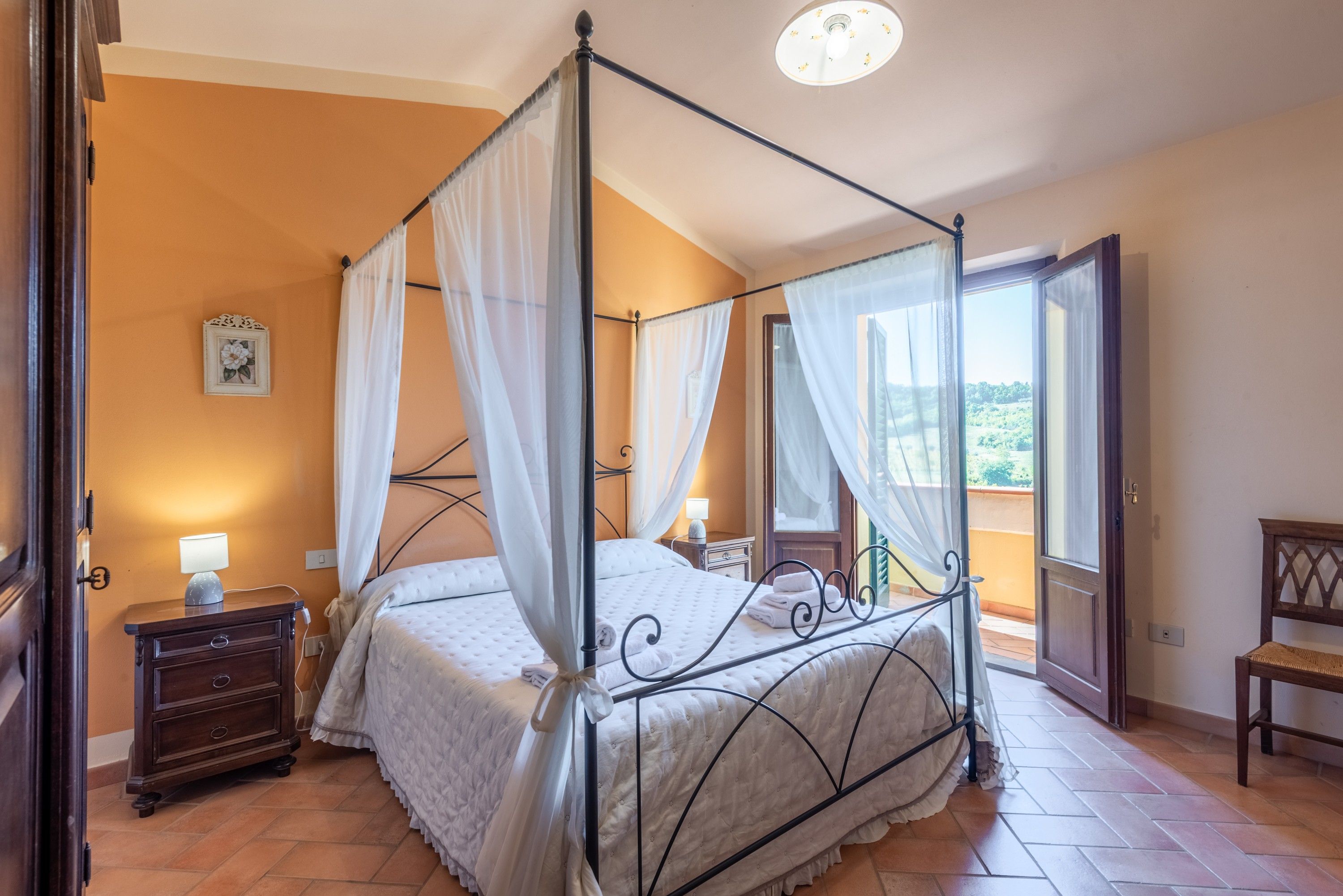 Un lit à baldaquin à quatre poteaux, recouvert d’une courtepointe blanche, une table de chevet avec une lampe, et une vue sur l’extérieur par une fenêtre.