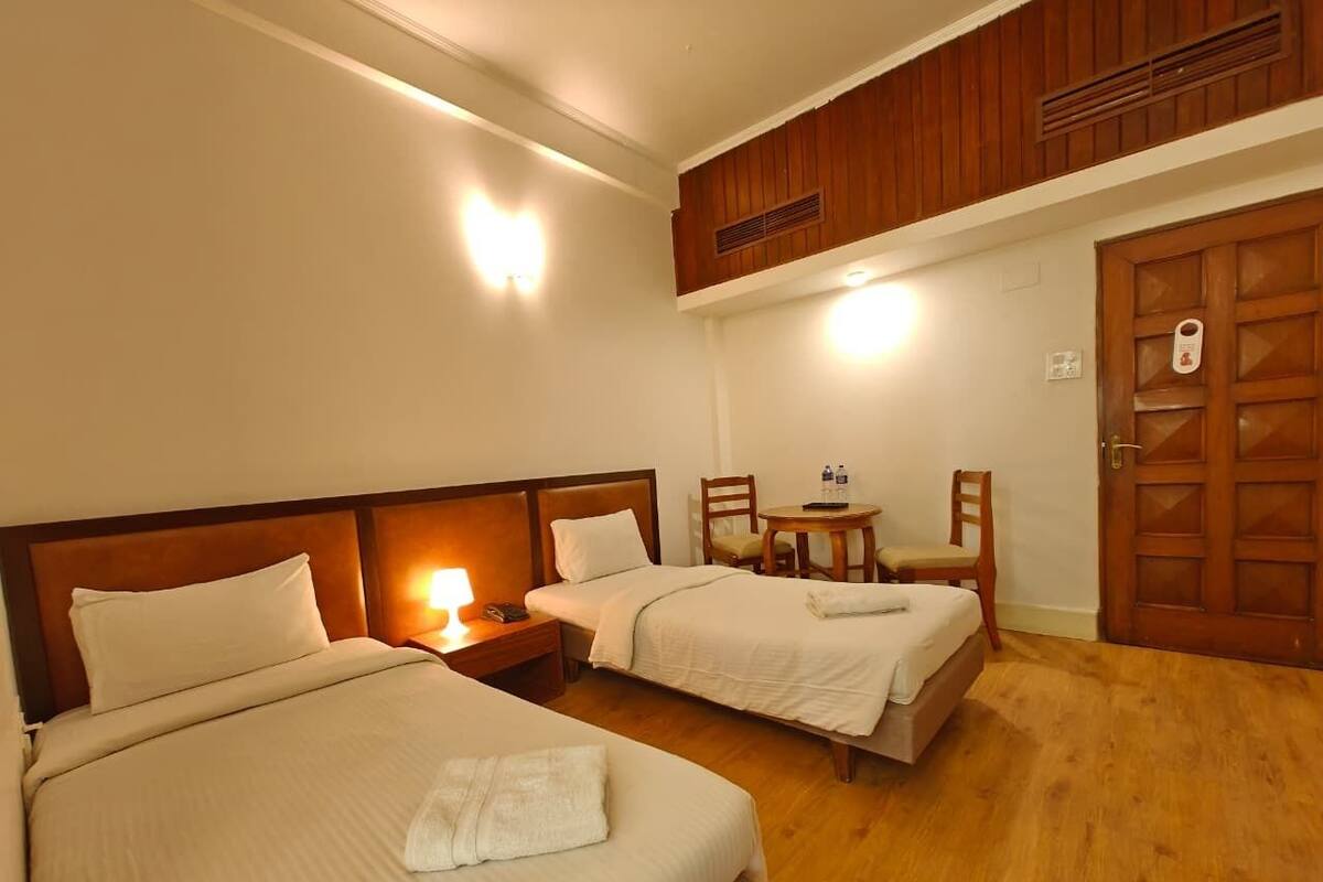 A hotel room with two beds, a small table with chairs, a wooden door, and a ceiling vent.