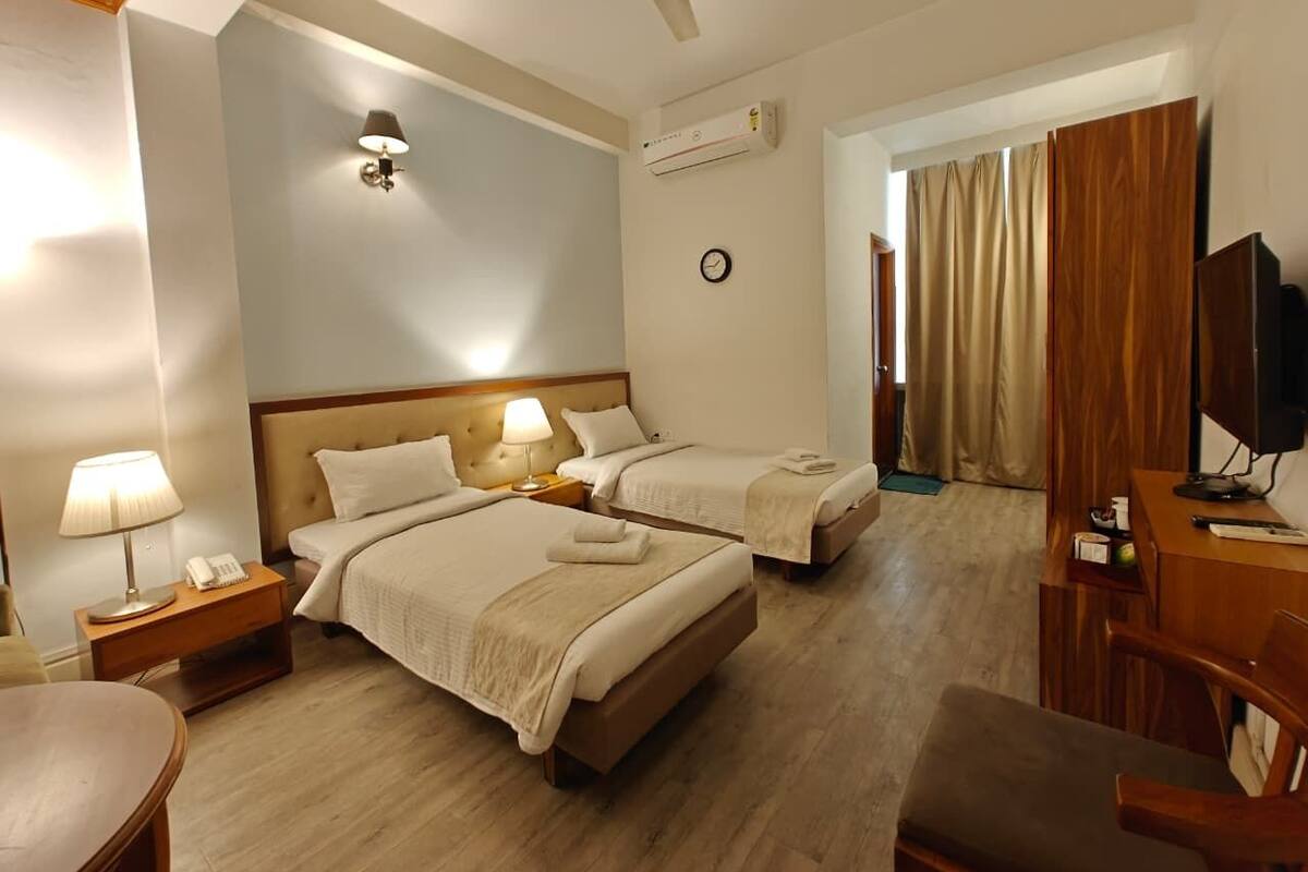 A hotel room with two beds, a desk, a chair, a TV, a lamp, a clock, and an air conditioning unit.
