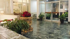 Un'ampia hall con pavimento in marmo, soffitto in legno e grandi finestre. Sono presenti piante in vaso e un'area salotto con tavoli e sedie.