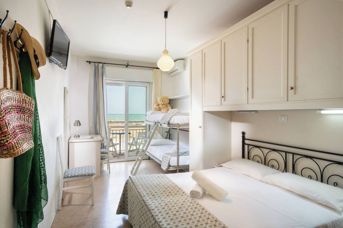 Een slaapkamer met stapelbedden, een klein bureau, een stoel, een televisie en een balkon met uitzicht.