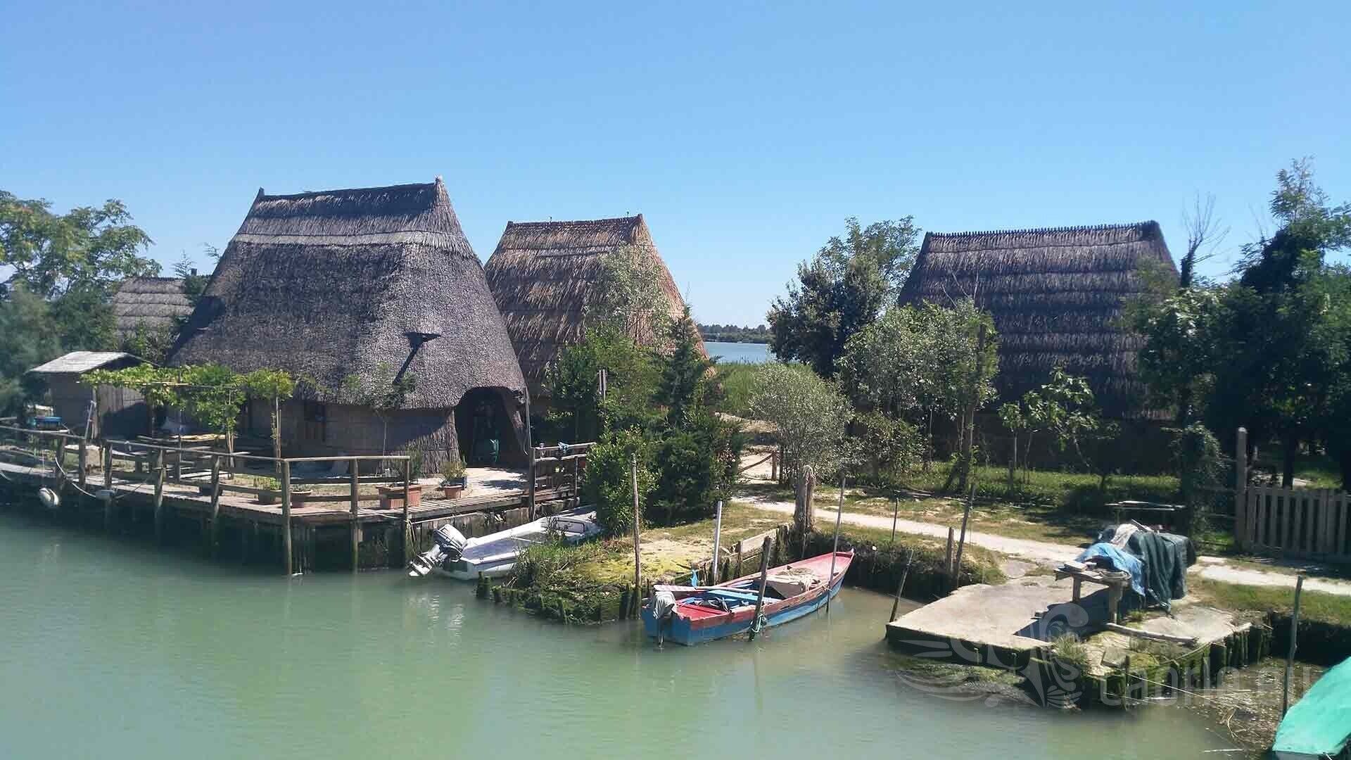 Thatched-roof huts by a calm water body, wooden docks, and a small boat.