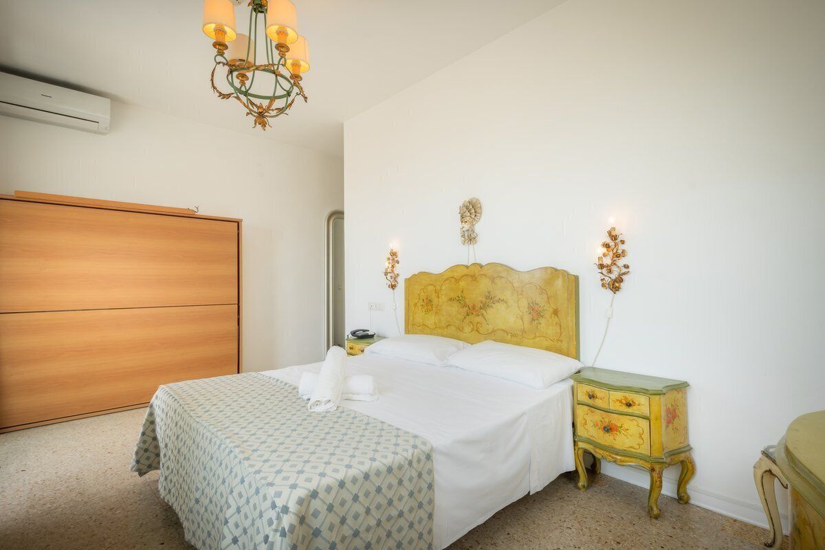 Ein ordentlich bezogenes Bett mit einem gemusterten Überwurf, ein Holzschrank, ein kleiner Nachttisch mit Lampe und eine an der Wand befestigte Uhr.