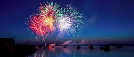 A nighttime scene with fireworks over a calm body of water, boats, and a building on the left.