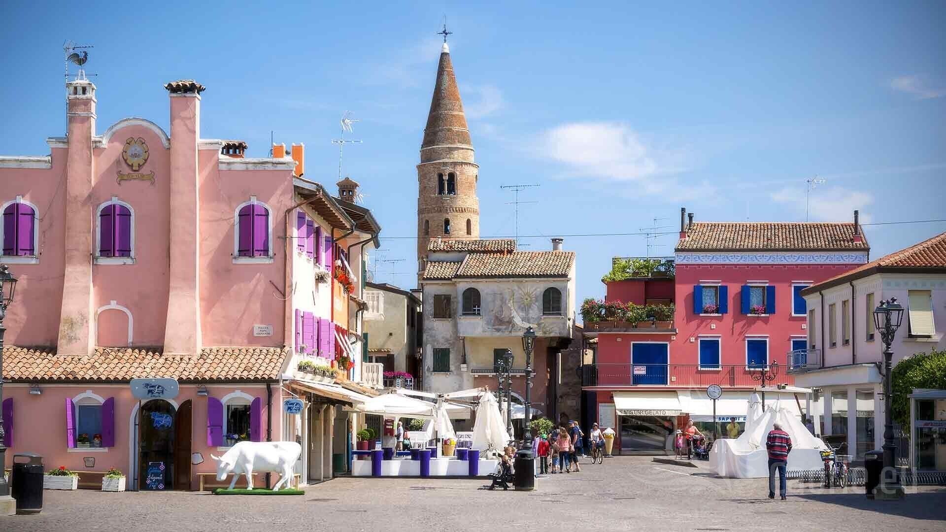 Una plaza histórica con edificios tradicionales, una torre de iglesia destacada y zonas con mesas al aire libre.