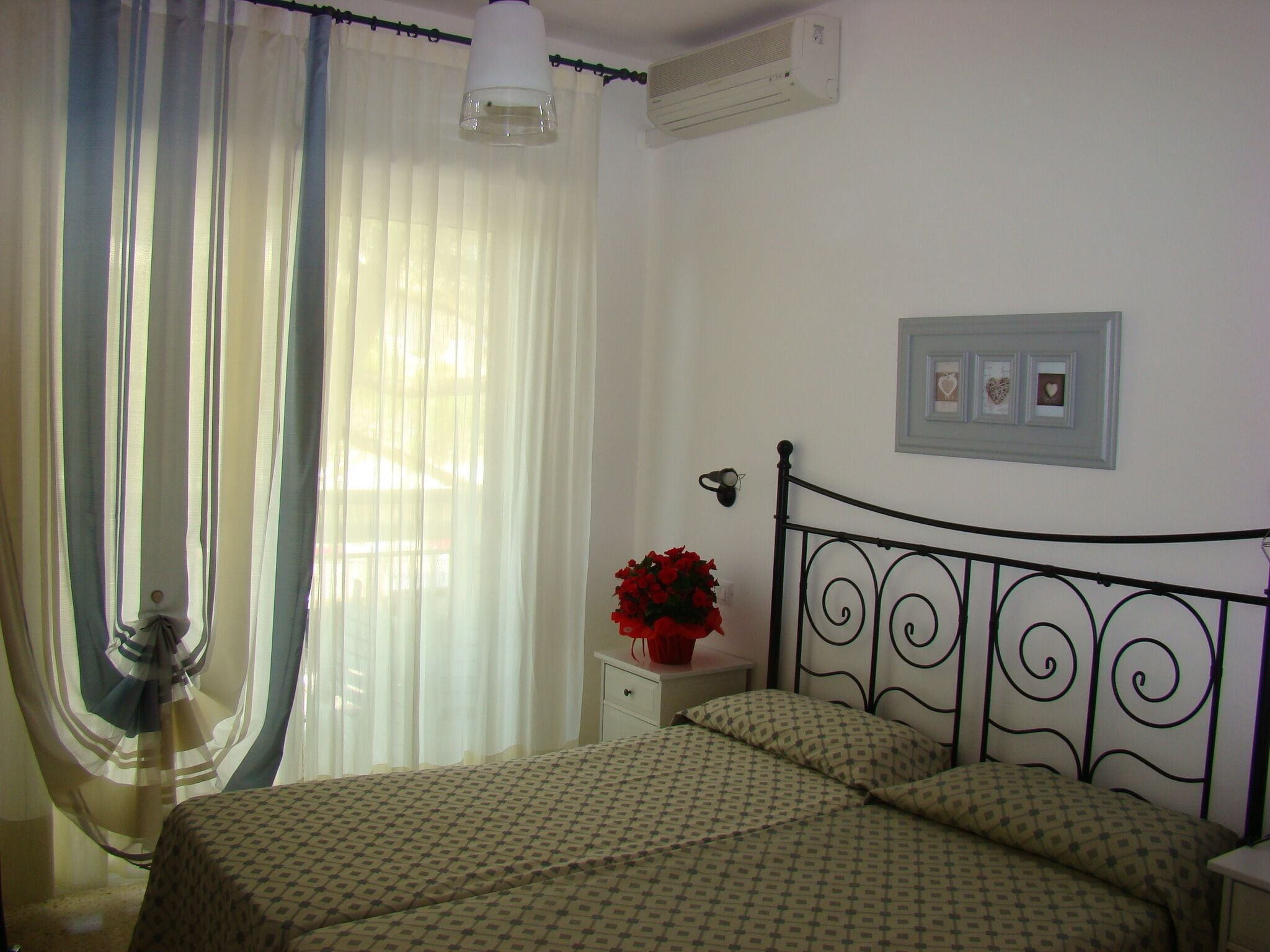 Un dormitorio con cama, cortinas, mesita de noche, aire acondicionado instalado en la pared y un cuadro enmarcado.