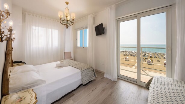 Une chambre d’hôtel avec un lit, une table de chevet, un lustre, une télévision et un balcon donnant sur la plage.