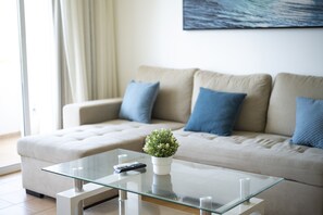 Ein modernes Wohnzimmer mit einem Glastisch, einem beige gefärbten Sofa mit blauen Kissen, einer kleinen Topfpflanze und einer Fernbedienung.