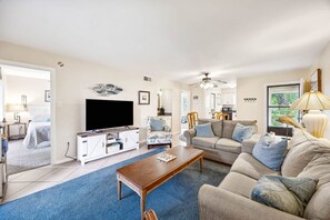 Ein geräumiges Wohnzimmer mit einem großen Flachbildfernseher, einem Holztisch, einem beigen Sofa und einem blauen Teppich.