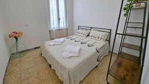 Un dormitorio ordenado con una cama, una mesita de noche con un florero y una ventana con cortinas translúcidas, además de una escalera metálica apoyada en la pared.
