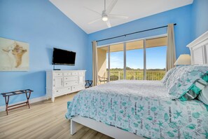 Ein Schlafzimmer mit Bett, Fernseher, weißem Schminktisch, Deckenventilator und großem Fenster mit Blick ins Freie.