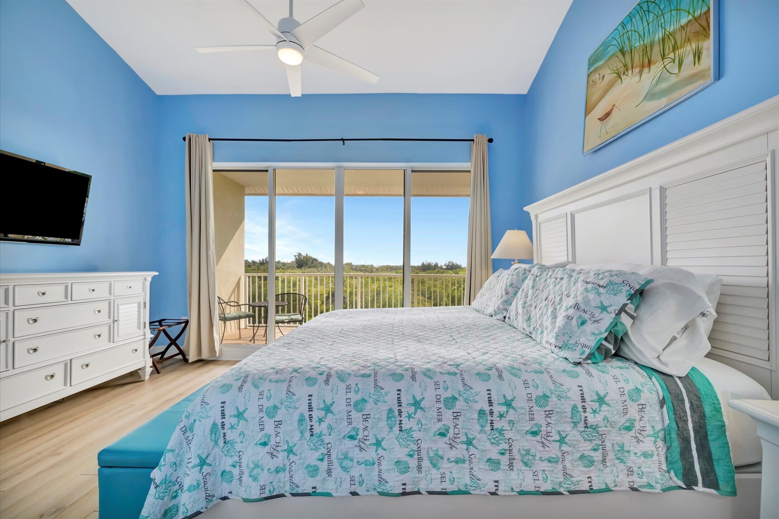 A bedroom with a large bed, a ceiling fan, a TV, a white dresser, and a balcony with a view of greenery.