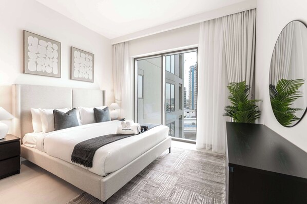Quarto de hotel moderno com uma cama grande, uma mesinha de cabeceira, um espelho e vista da cidade por grandes janelas.