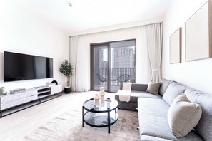 Uma sala de estar moderna com uma televisão de tela plana grande, um sofá cinza, uma mesa de centro de vidro e uma varanda com vista para edifícios altos.