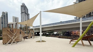 Un espacio al aire libre con arena, que incluye un parque infantil con columpios y estructuras para escalar, todo bajo una gran pérgola.
